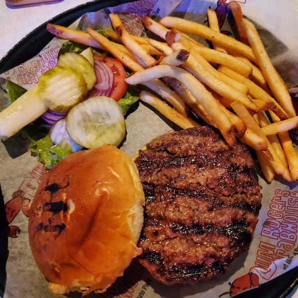 A grilled burger patty and bun with fries, pickles, and red onion on a branded paper liner.