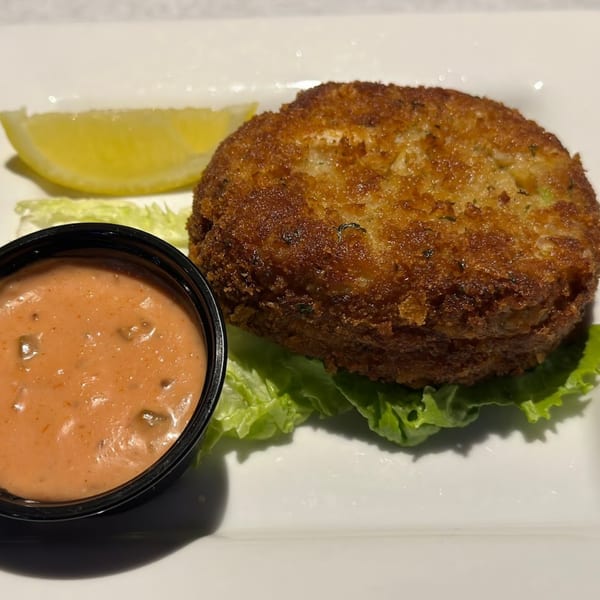 A golden-brown crab cake on lettuce with a lemon wedge and a side of pink sauce.