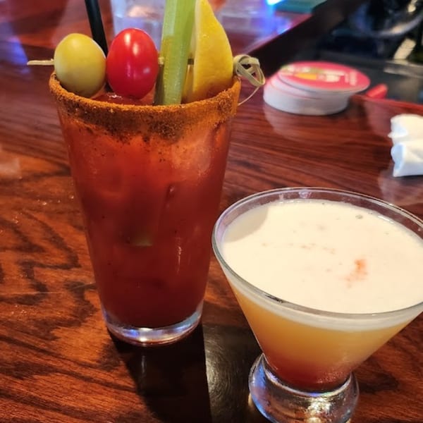 A Bloody Mary garnished with celery, lemon, olive, and tomato, next to a foamy orange cocktail.