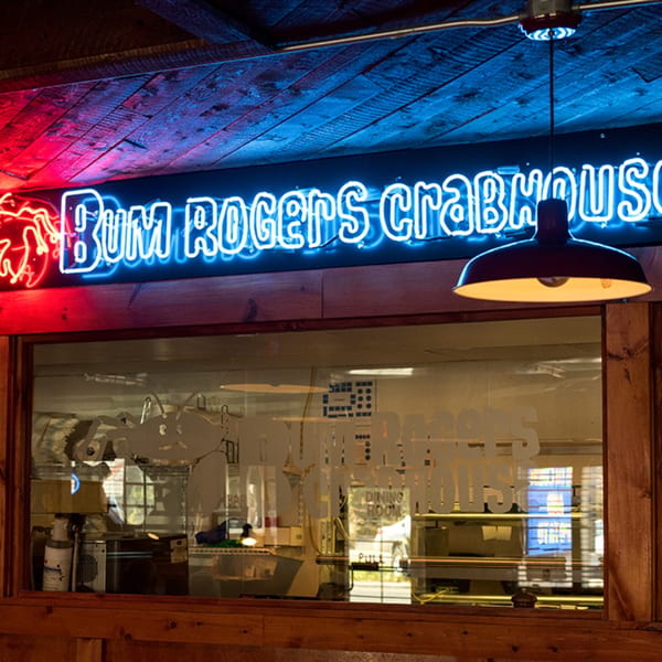 Neon sign for Bum Rogers Crabhouse in blue and red, above a window with frosted lettering.